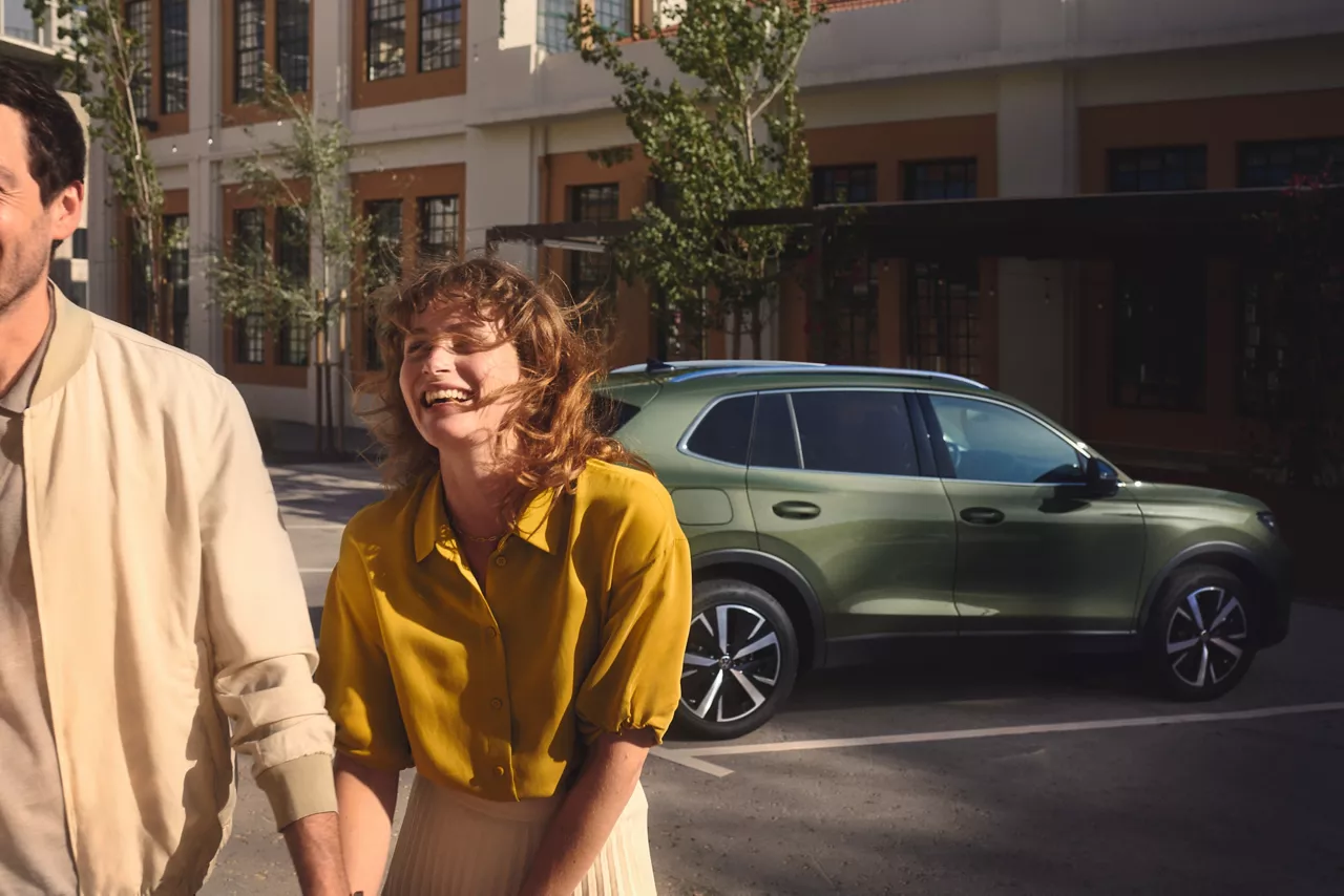 Uśmiechnięta kobieta idzie za rękę z mężczyzną. Za nimi stoi zaparkowany, zielony samochód z Grupy Volkswagen.